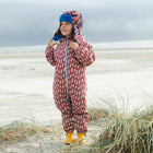 A child on a beach in an EcoSplash waterproof pink puddle suit with lightning bolts, wearing a hat and yellow wellies.